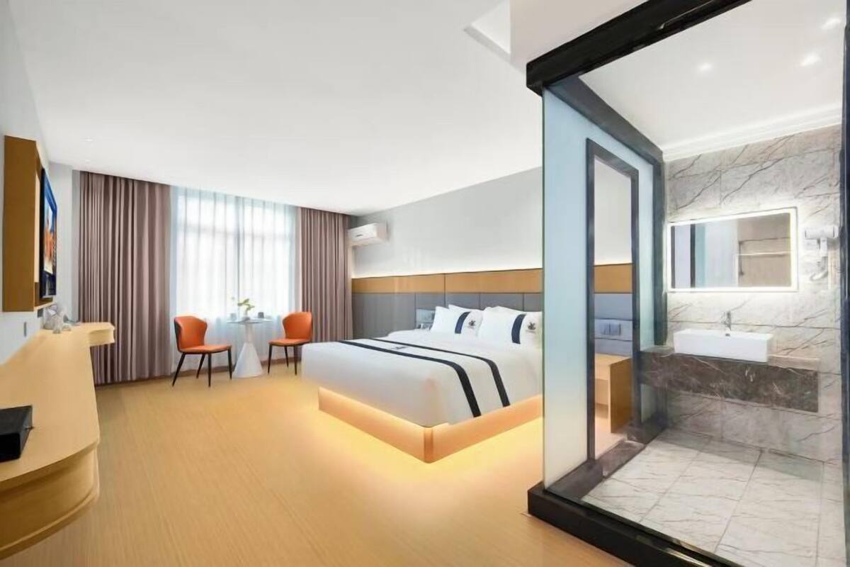 Habitación de hotel con cama, escritorio, dos sillas, una mesita, baño con lavabo y espejo, y un separador de vidrio.