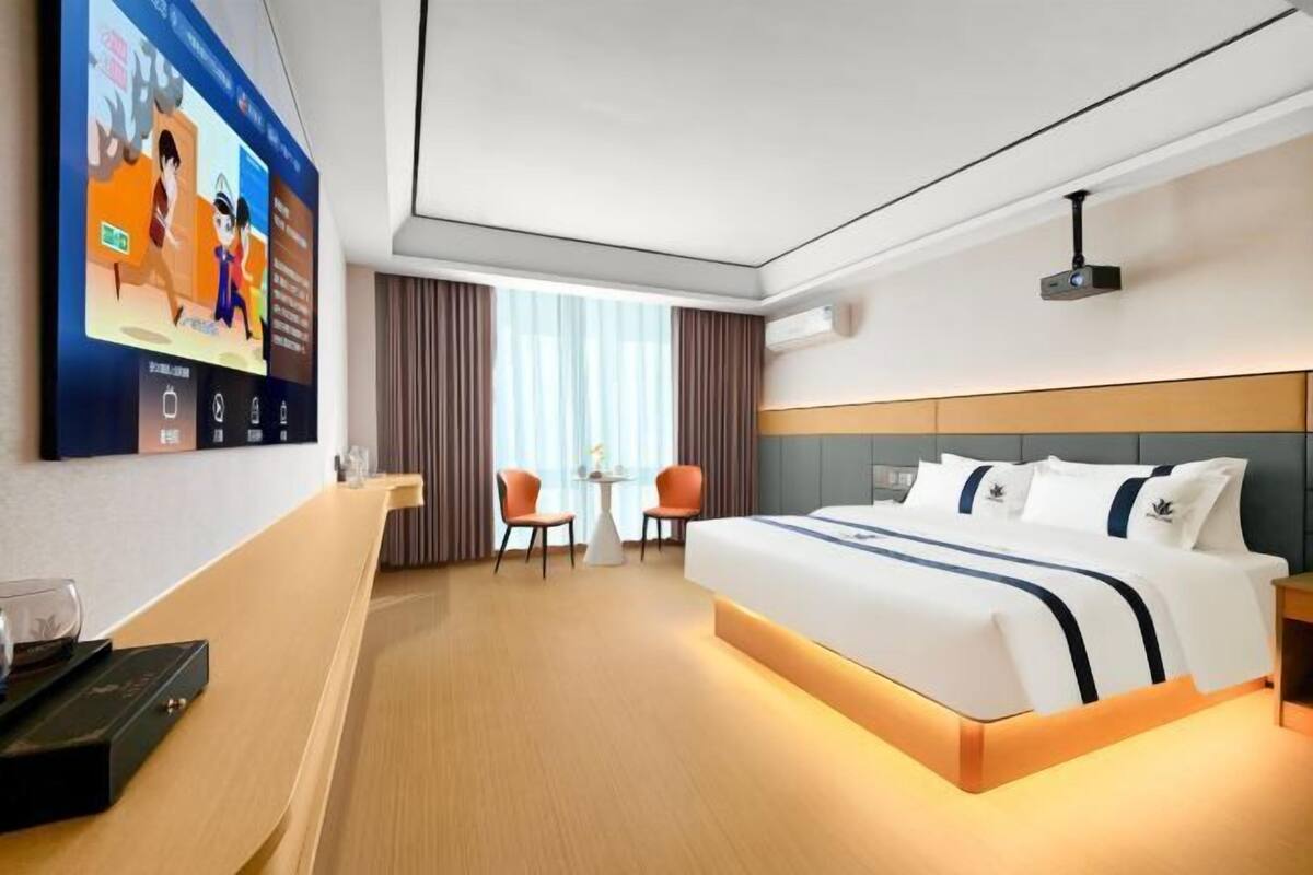Una habitación de hotel con cama, televisión montada en la pared, escritorio con silla y ventana con cortinas.
