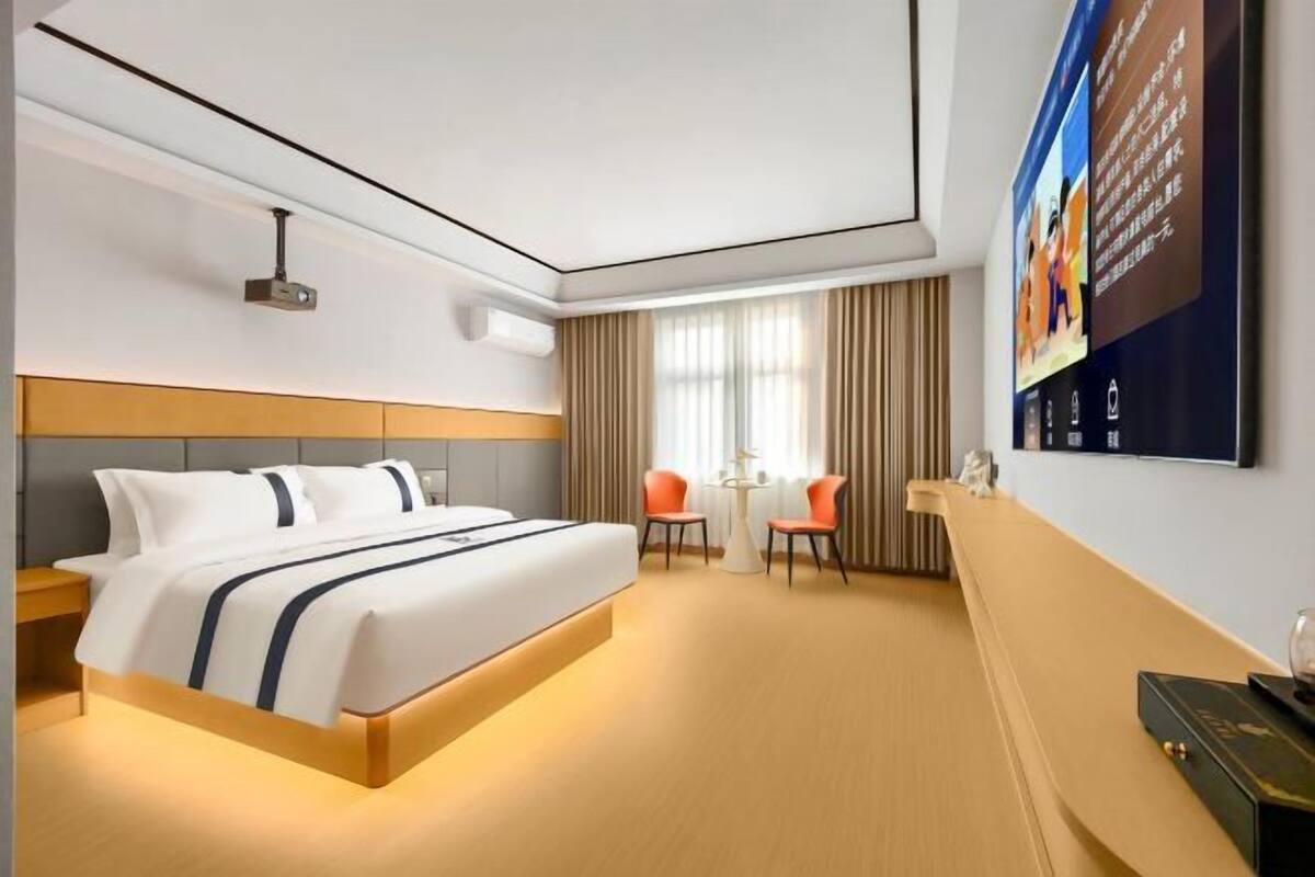 Una habitación de hotel con cama, mesita de noche, sillas, televisor y una pantalla montada en la pared.