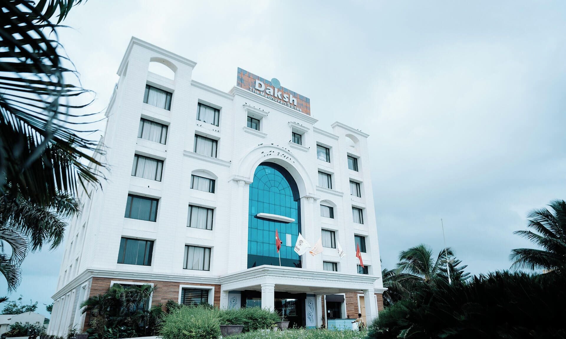 A white building with a blue entrance, palm trees, and flags.
