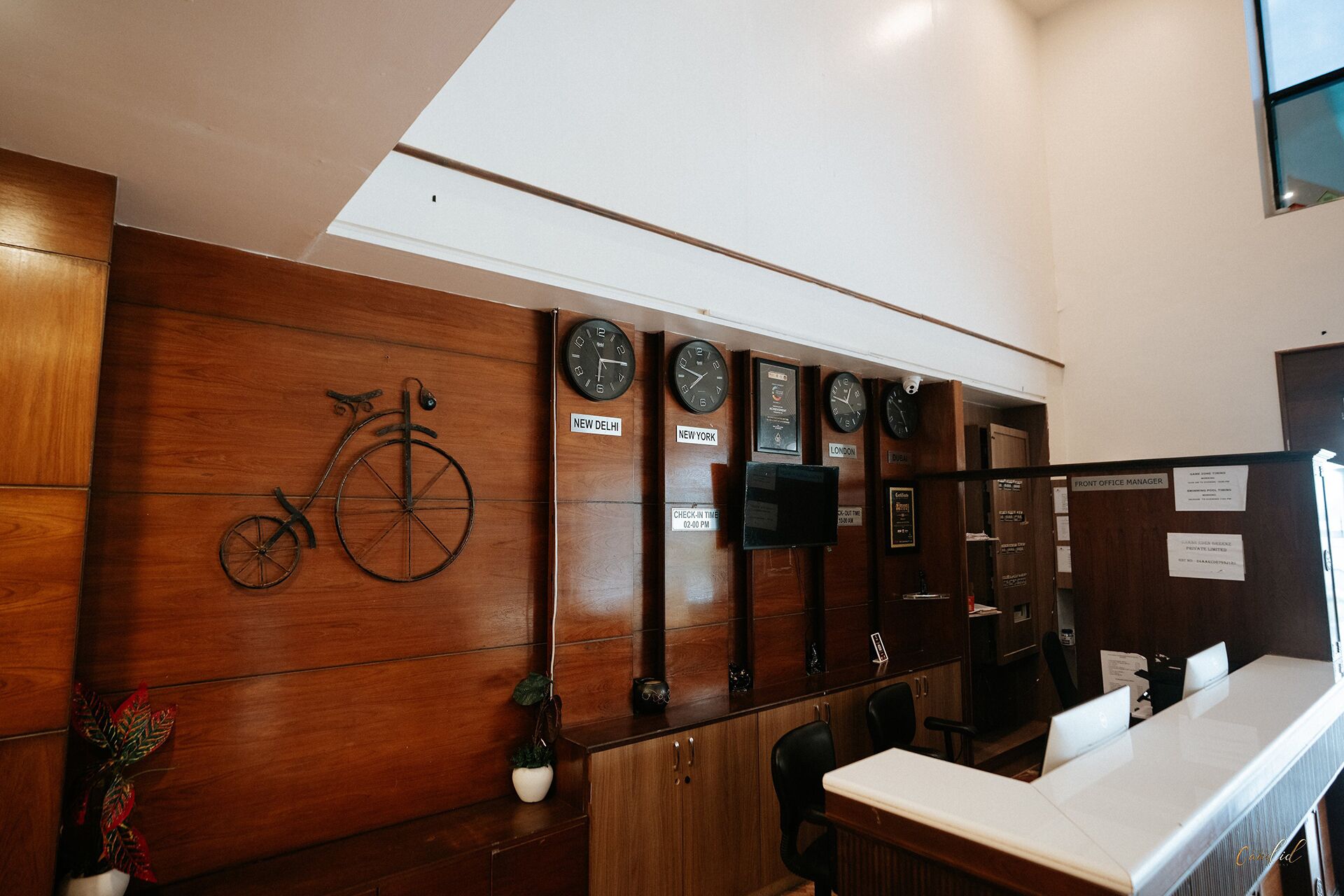 Bancada de recepção com parede de painéis de madeira, relógios exibindo diferentes fusos horários e uma bicicleta decorativa.