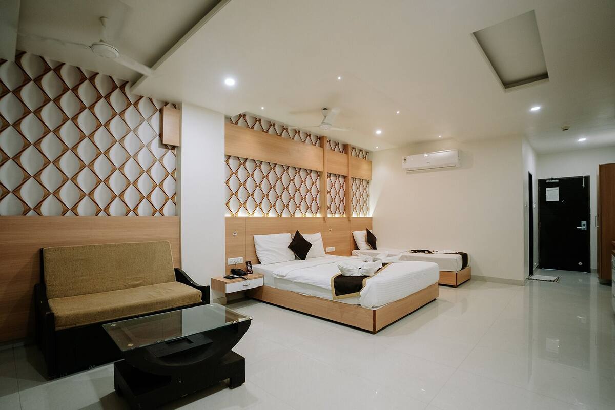 A modern hotel room with a sofa, a glass coffee table, a bed with a wooden headboard, and a decorative wall panel.