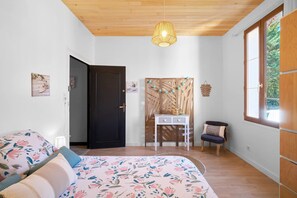 A bedroom with a bed, a desk, a chair, a hanging light fixture, and a window.