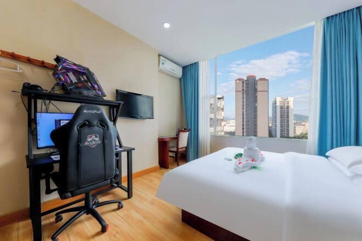 A hotel room with a bed, a desk with a computer and a gaming chair, a TV, and a window with a city view.