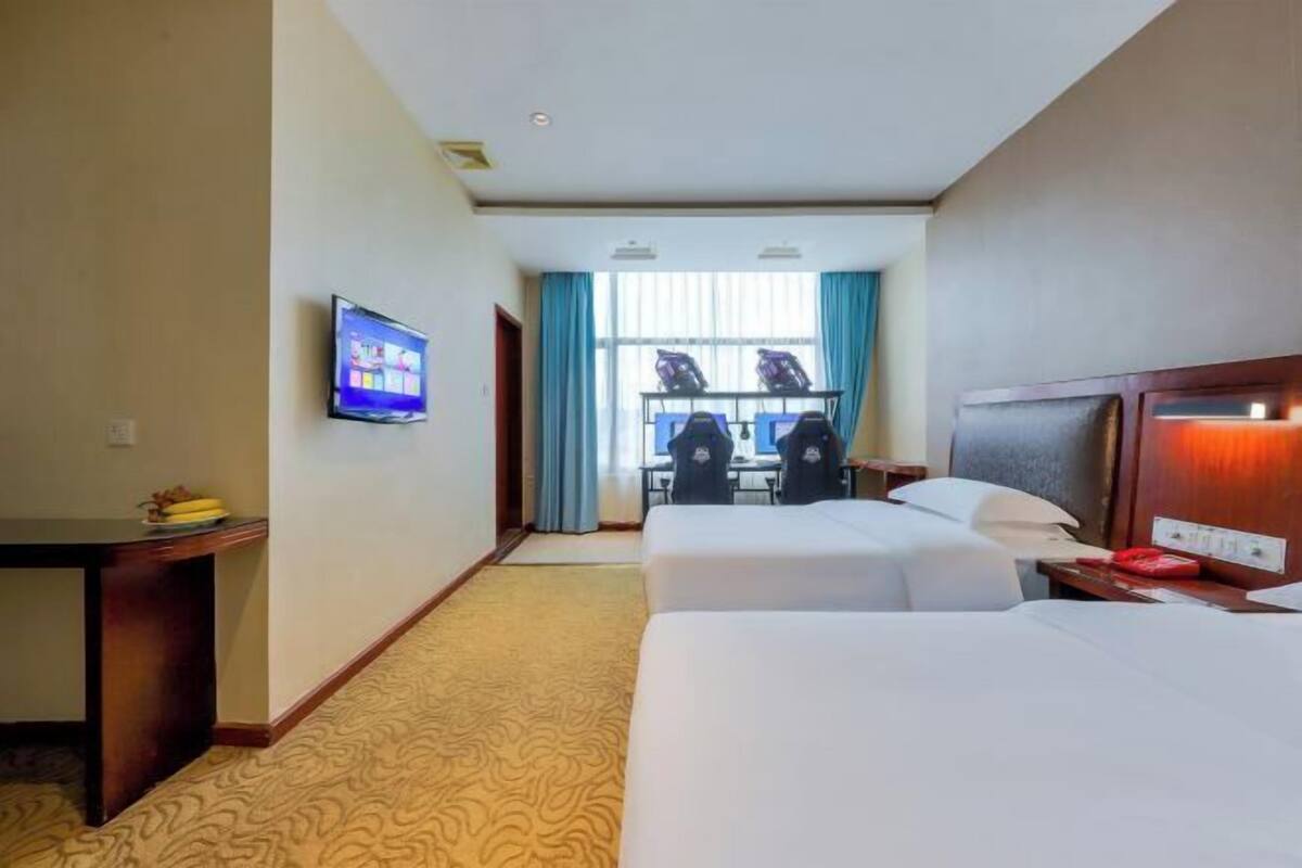 A hotel room with two beds, a desk, a television, and a view of the city through large windows.