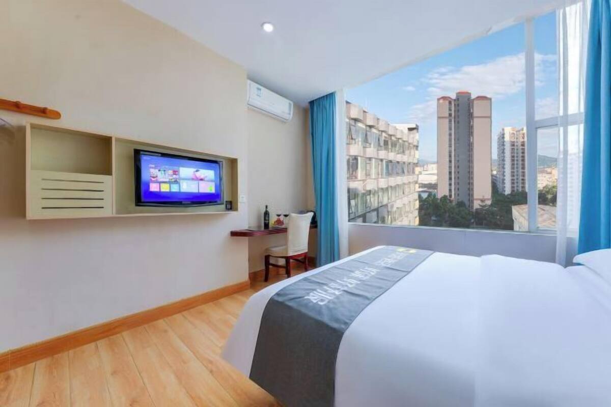 A hotel room with a bed, a TV, a desk, and a window with a city view.