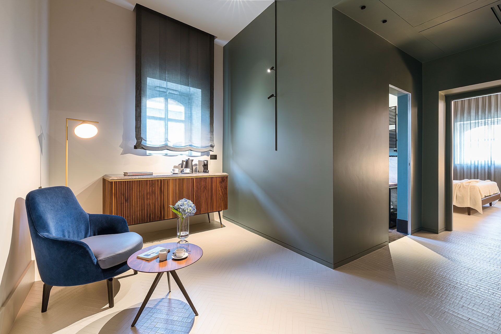 Ein modernes Hotelzimmer mit blauem Sessel, einem kleinen runden Tisch mit einer Vase und Blick auf ein Schlafzimmer mit Bett und Fenster.