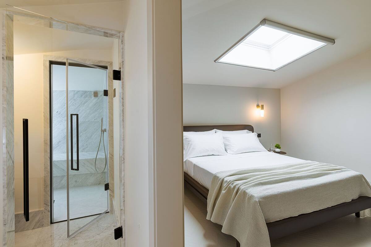 Une chambre d’hôtel moderne dotée d’une douche en verre, d’un lit avec du linge de lit blanc et d’une lucarne au-dessus du lit.