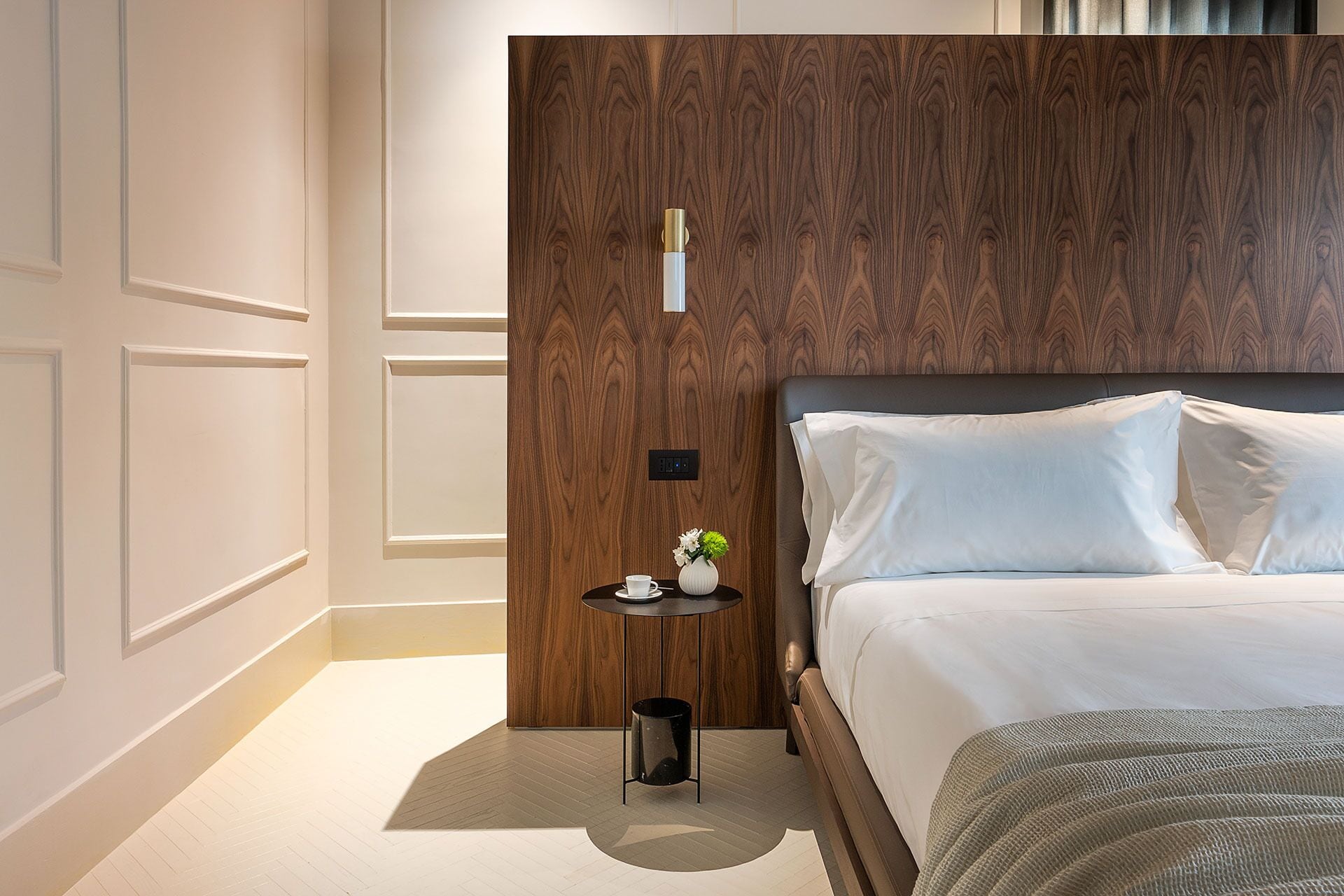 A modern hotel room with a wooden headboard, a small bedside table with a vase and a cup, and a neatly made bed.