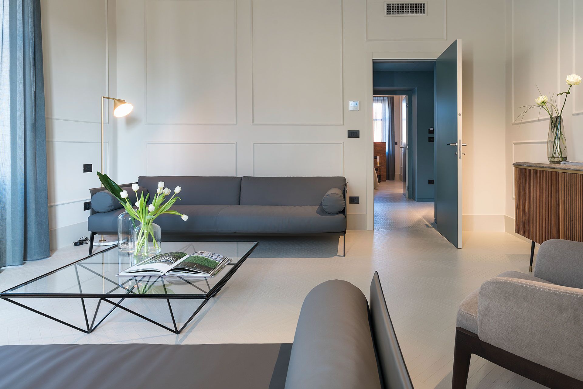 Una sala de estar moderna con un sofá gris, una mesa de centro de vidrio con revistas, y un jarrón con flores blancas.