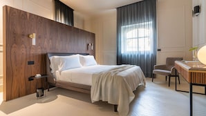 A modern hotel room with a large bed, a desk with a lamp, a chair, and a large window with curtains. -  Byssus Suites (Syracuse)