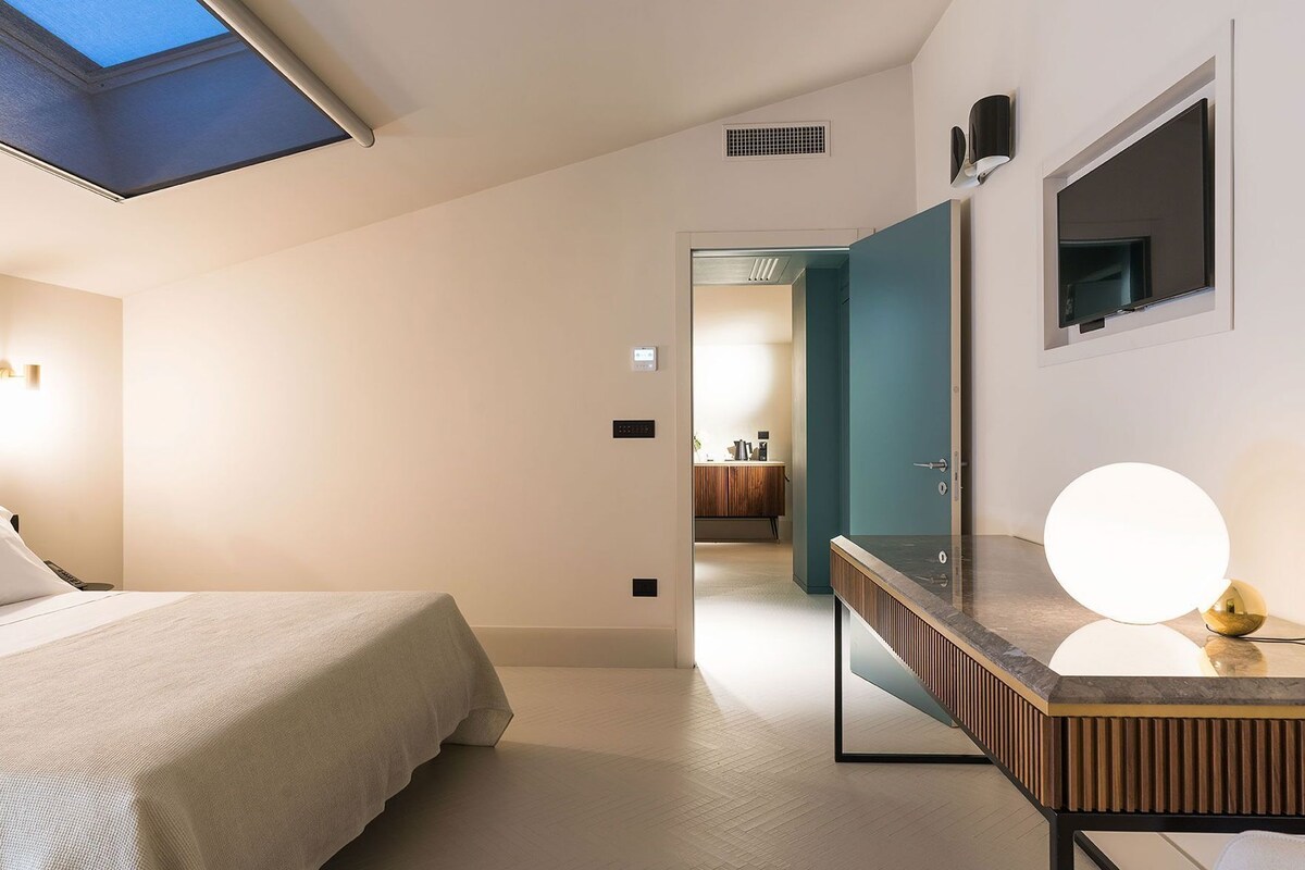 A bedroom with a skylight, a bedside table, a bed, a bathroom with a mirror, a sink, and a round light fixture.