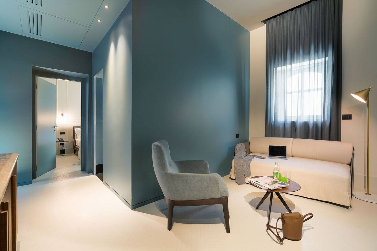 Un salon moderne avec un fauteuil gris, un canapé, une petite table ronde sur laquelle se trouvent un ordinateur portable et un magazine, et un sac à main marron posé sur le sol.