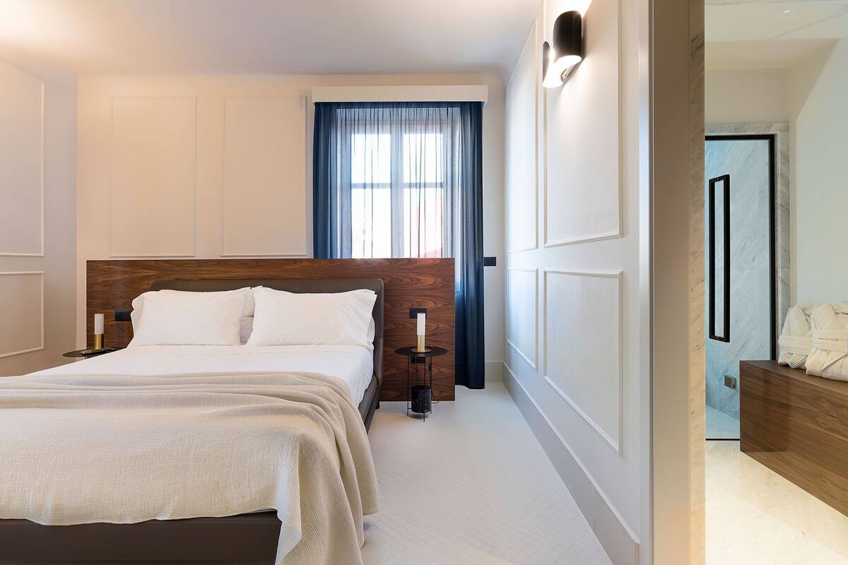 Une chambre à coucher avec un grand lit, une tête de lit en bois, une table de chevet avec une lampe et une fenêtre avec des rideaux.