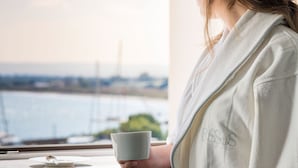 A person holding a white mug by a window with a view of a body of water and distant structures. -  Byssus Suites (Syracuse)