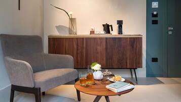 Une chambre d’hôtel moderne avec un fauteuil gris, un meuble en bois orné d’un vase de fleurs, une machine à café et une table ronde sur laquelle se trouvent un livre et des lunettes.