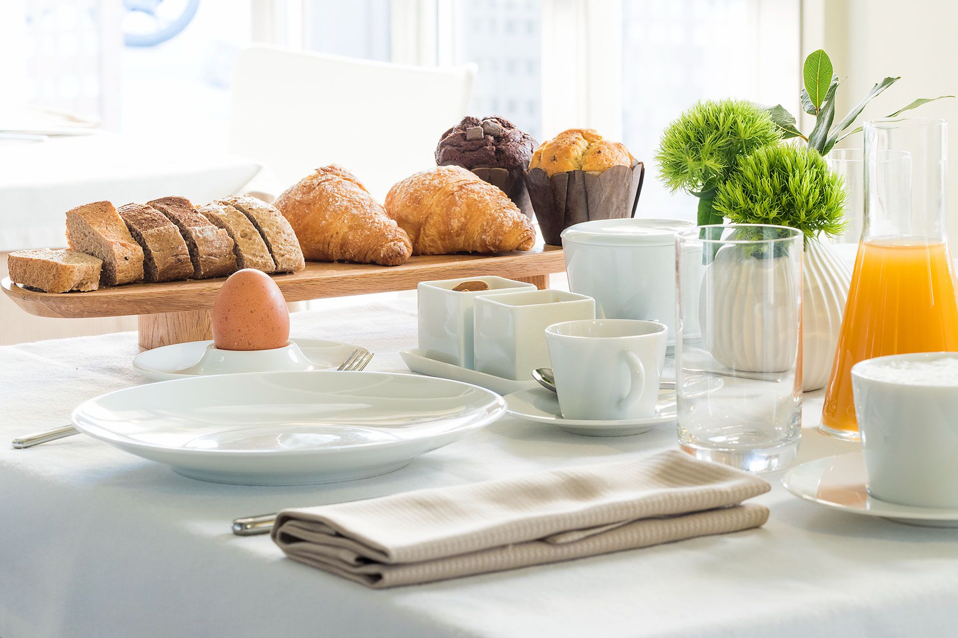 Desayuno con pan, bollería, tazas de café y un vaso de zumo de naranja en una mesa.