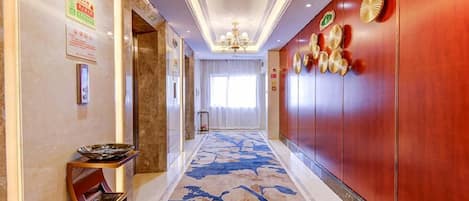 A hotel corridor with a patterned carpet, a reception desk, a chandelier, and a wall with decorative plates.
