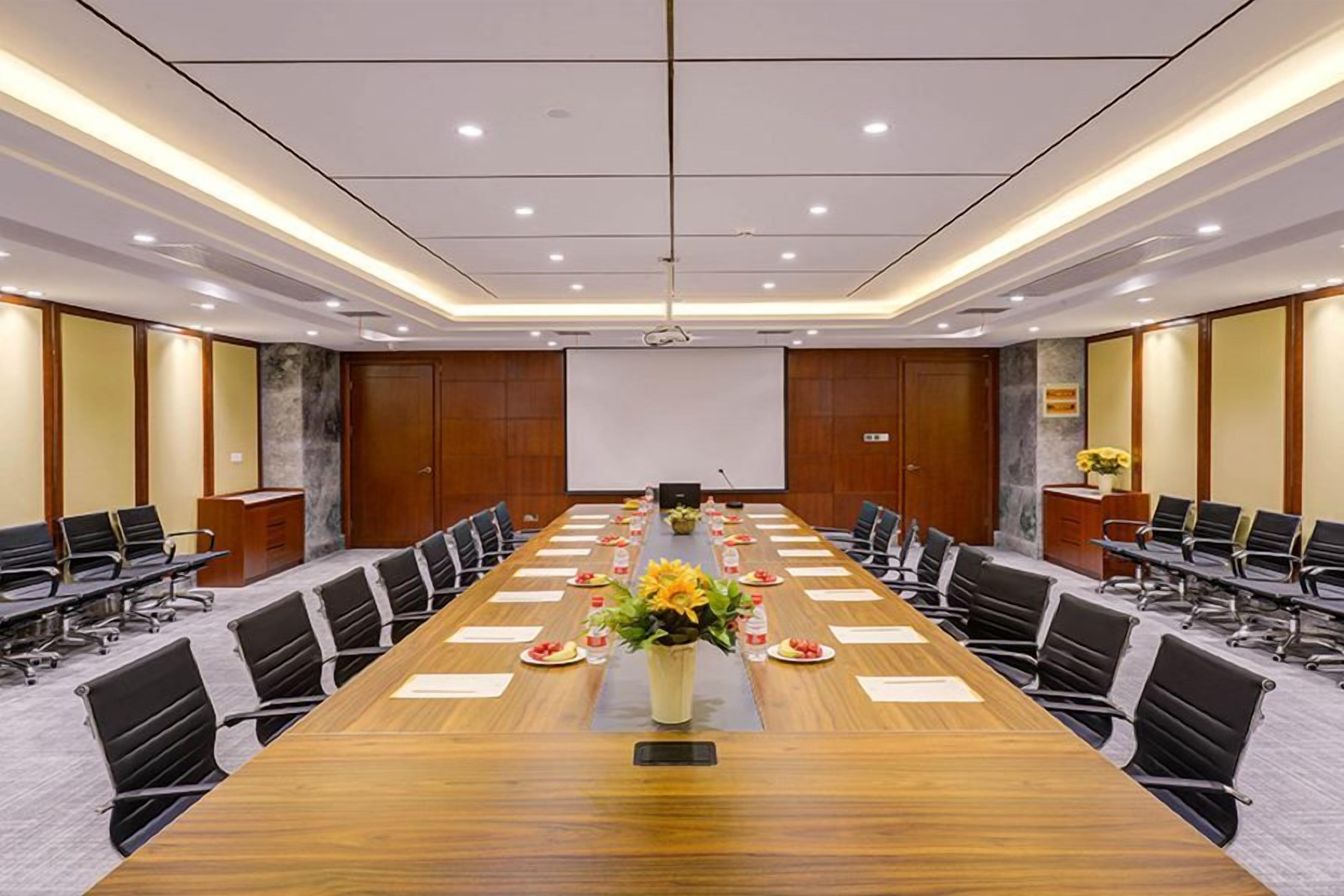 Un salón de conferencias con una larga mesa, sillas, una pantalla de proyector y un centro de mesa floral.