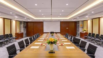 Un salón de conferencias con una larga mesa, sillas, una pantalla de proyector y un centro de mesa floral.