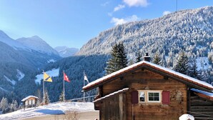 Eine Holzhütte mit geneigtem Dach, umgeben von schneebedeckten Bergen und blauem Himmel.