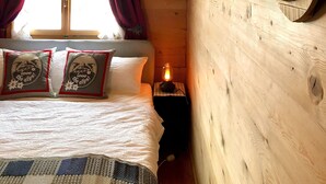 Ein Holzhütten-Zimmer mit einem Bett, zwei Kissen, einem Nachttisch mit einer Laterne und einem Fenster mit Vorhängen.