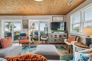 A spacious living room with a wooden ceiling, large windows, a television, and a view of the outdoors. - Lighthouse Cottage! Pet Friendly, Ocean View w/Screen Porch, Beach Access Next to Property (Folly Beach)