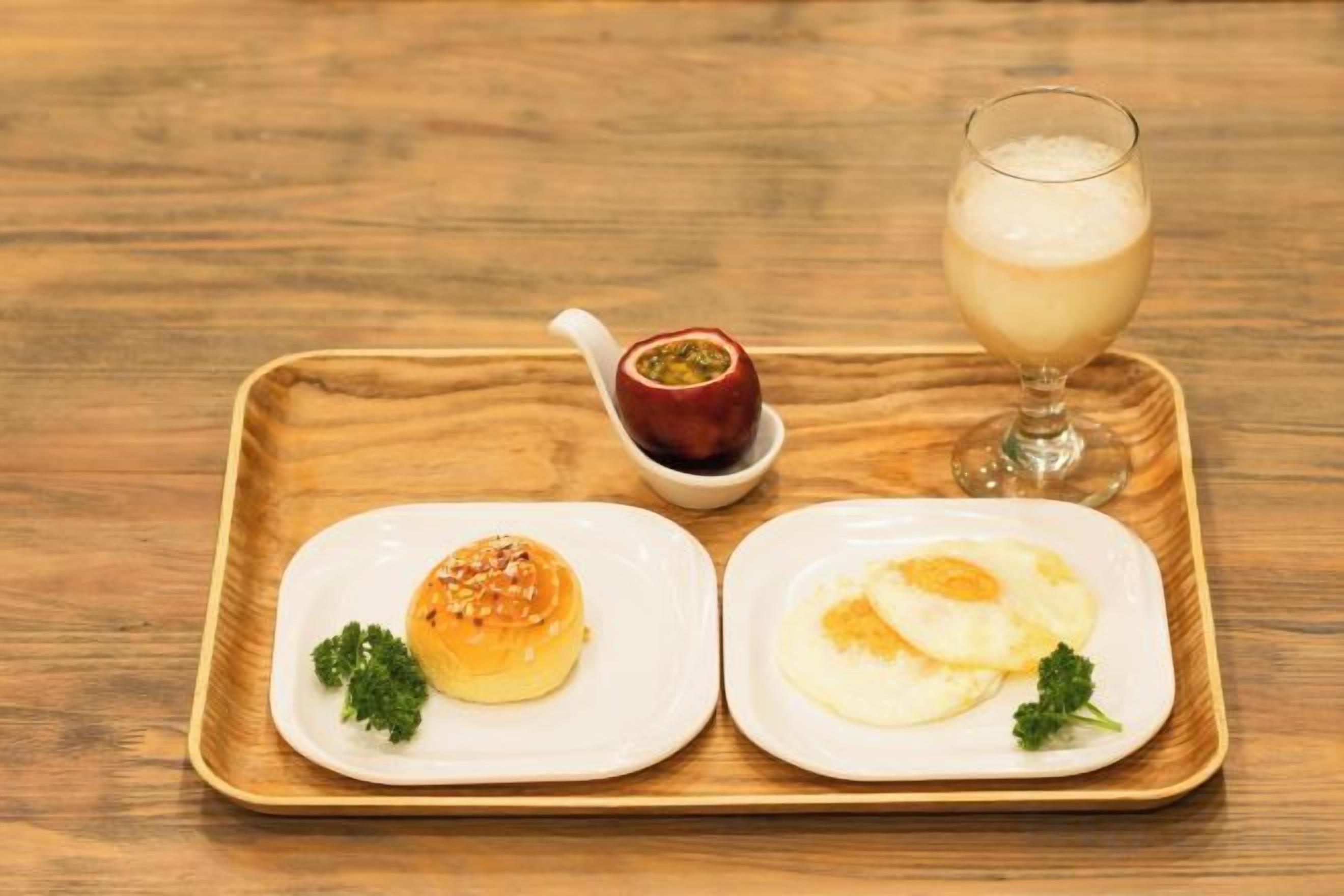 A tray with two plates of food, one with a fried egg and the other with a bun, and a glass of a creamy beverage on a wooden table.