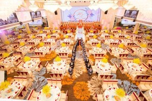 Un salón de banquetes con filas de mesas y sillas, un escenario con fondo decorativo, y una mesa larga con pasillo central.