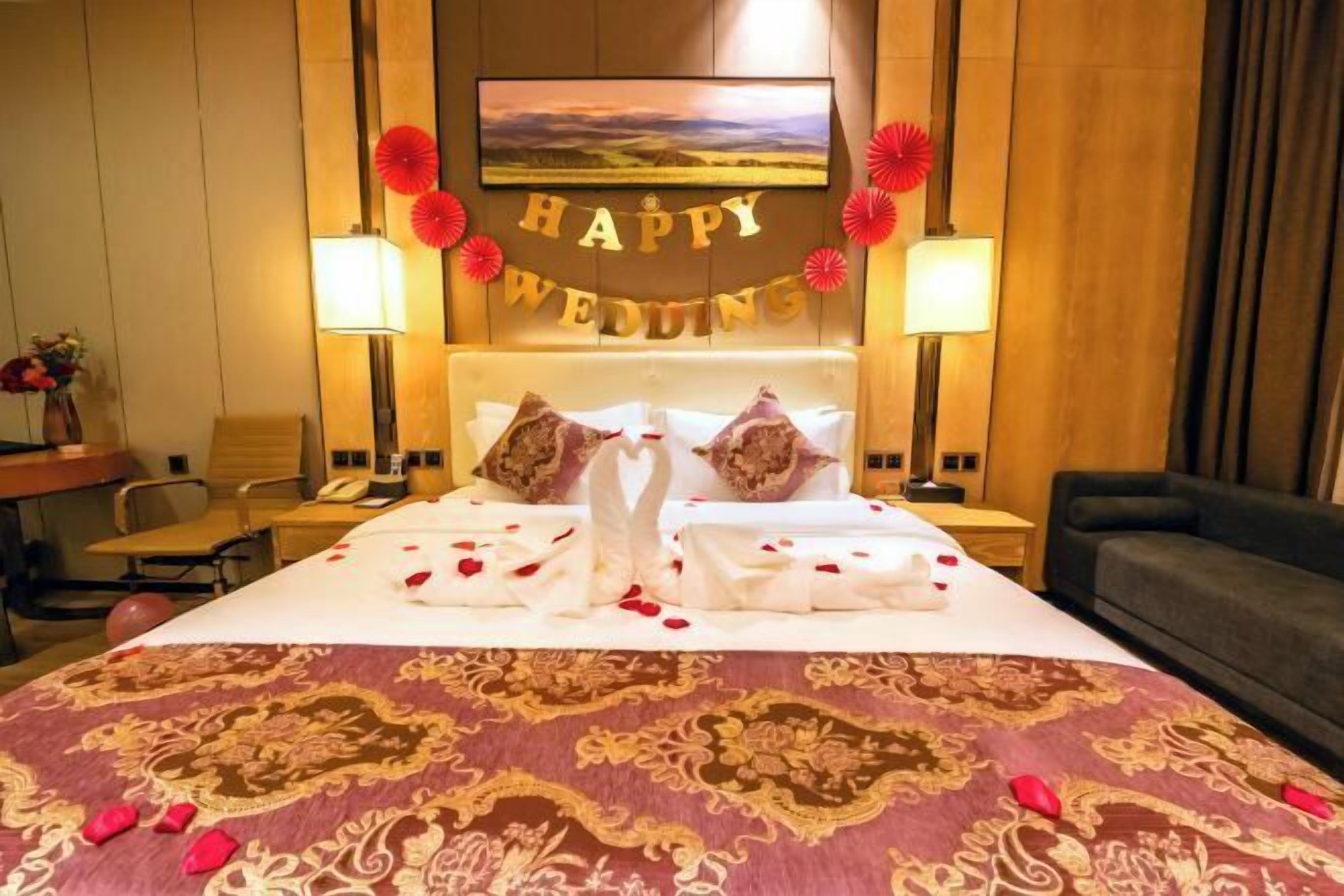 A hotel room with a bed, a 'HAPPY WEDDING' sign, a TV, and a lamp.