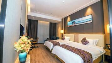 A hotel room with two beds, a desk, a chair, a vase of flowers, and a painting on the wall.