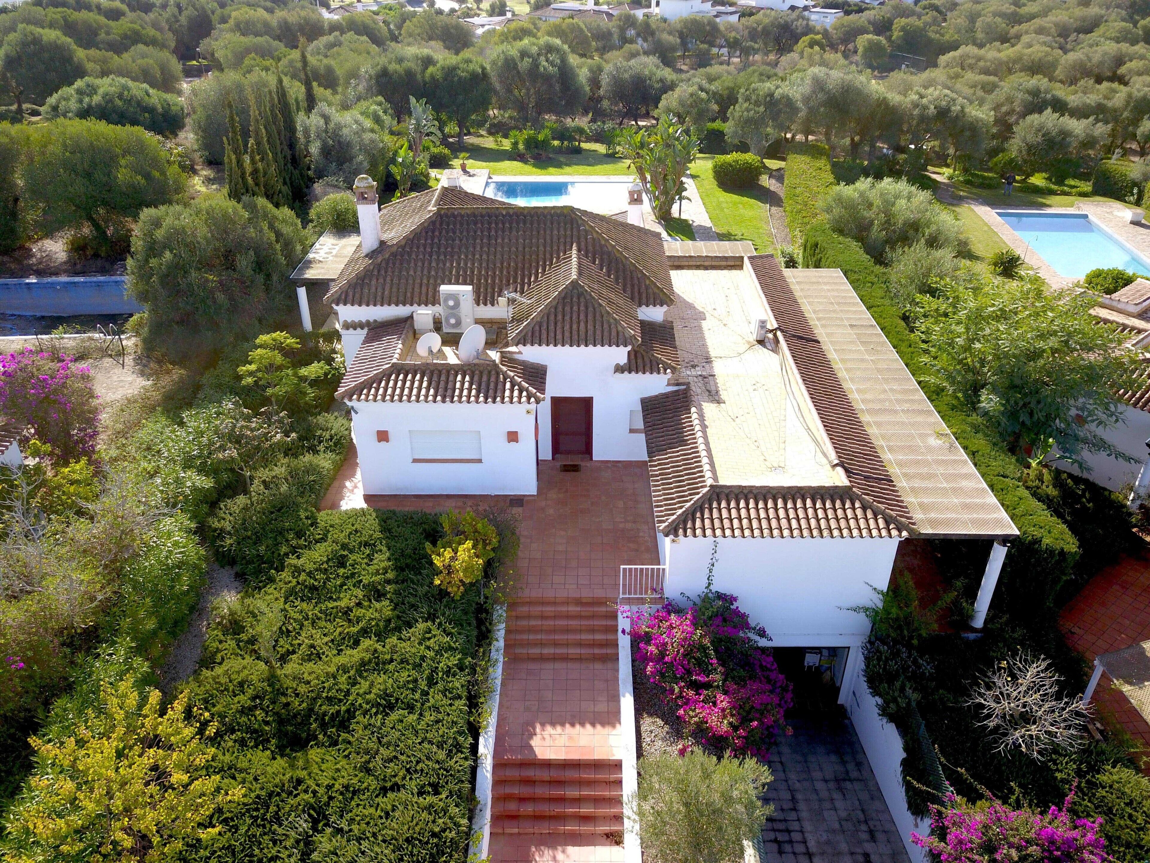Una villa blanca con techo de tejas, rodeada de exuberante vegetación y una piscina.