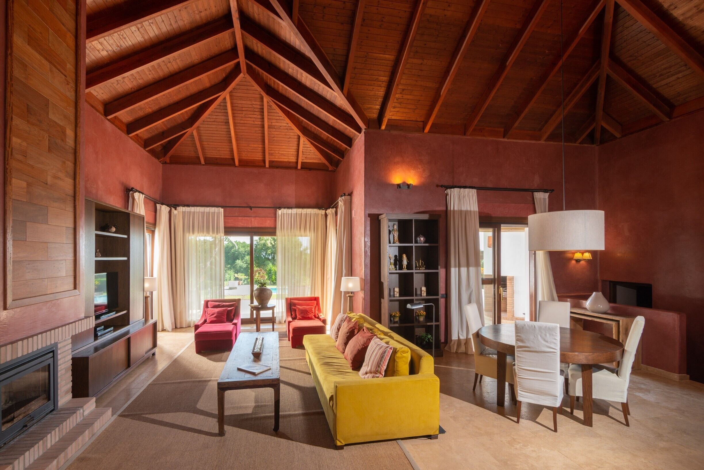 Een ruime woonkamer met een hoge plafond, een open haard, een gele bank, een eettafel met stoelen en een boekenkast.