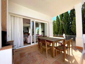 Un balcón con mesa y sillas de madera, una puerta corrediza de vidrio que da a una habitación interior, y vistas a árboles.
