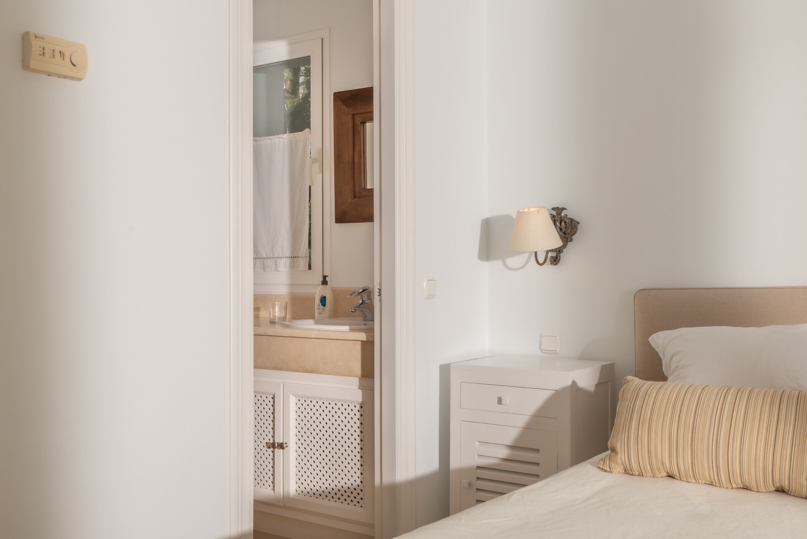 Ein Schlafzimmer mit Bett, Nachttisch, Wandlampe, Badezimmer mit Waschbecken und Spiegel sowie Thermostat an der Wand.
