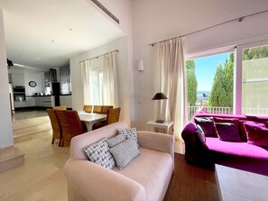 Una sala de estar moderna con un sofá, un comedor con sillas, una cocina al fondo y ventanas grandes con cortinas.