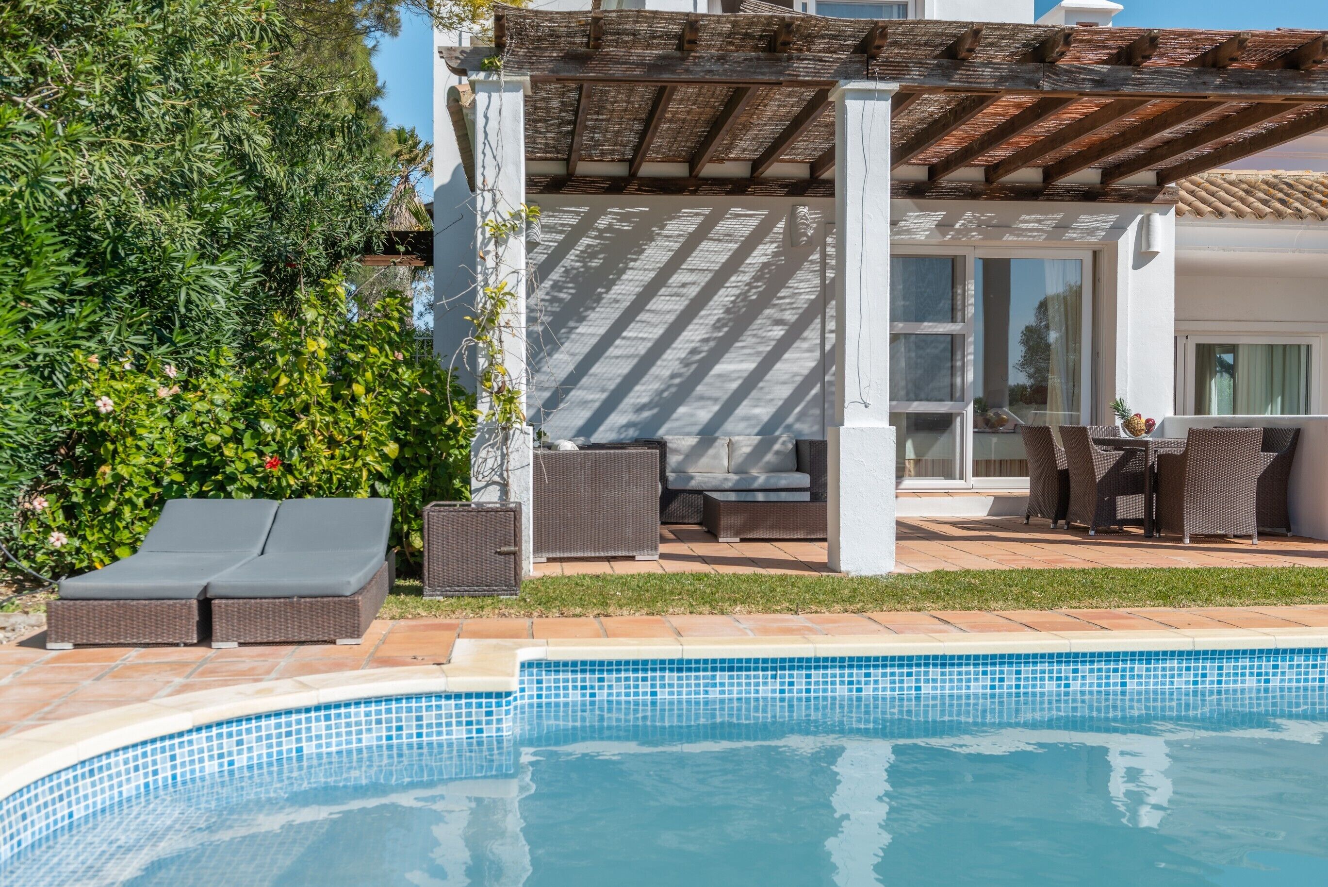 Une piscine avec des chaises longues, un patio couvert avec des meubles d’extérieur et une vue sur une maison dotée de grandes fenêtres.