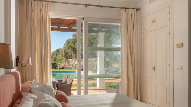 Ein Schlafzimmer mit Bett, Nachttisch, Lampe sowie Blick auf einen Swimmingpool und einer Glasschiebetür, die zu einem Außenbereich führt.