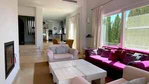 Ein modernes Wohnzimmer mit einem weißen Sofa, einem rosafarbenen Sofa und einem weißen Couchtisch. Im Hintergrund befindet sich eine Küche, und ein Balkon bietet Blick auf Bäume.