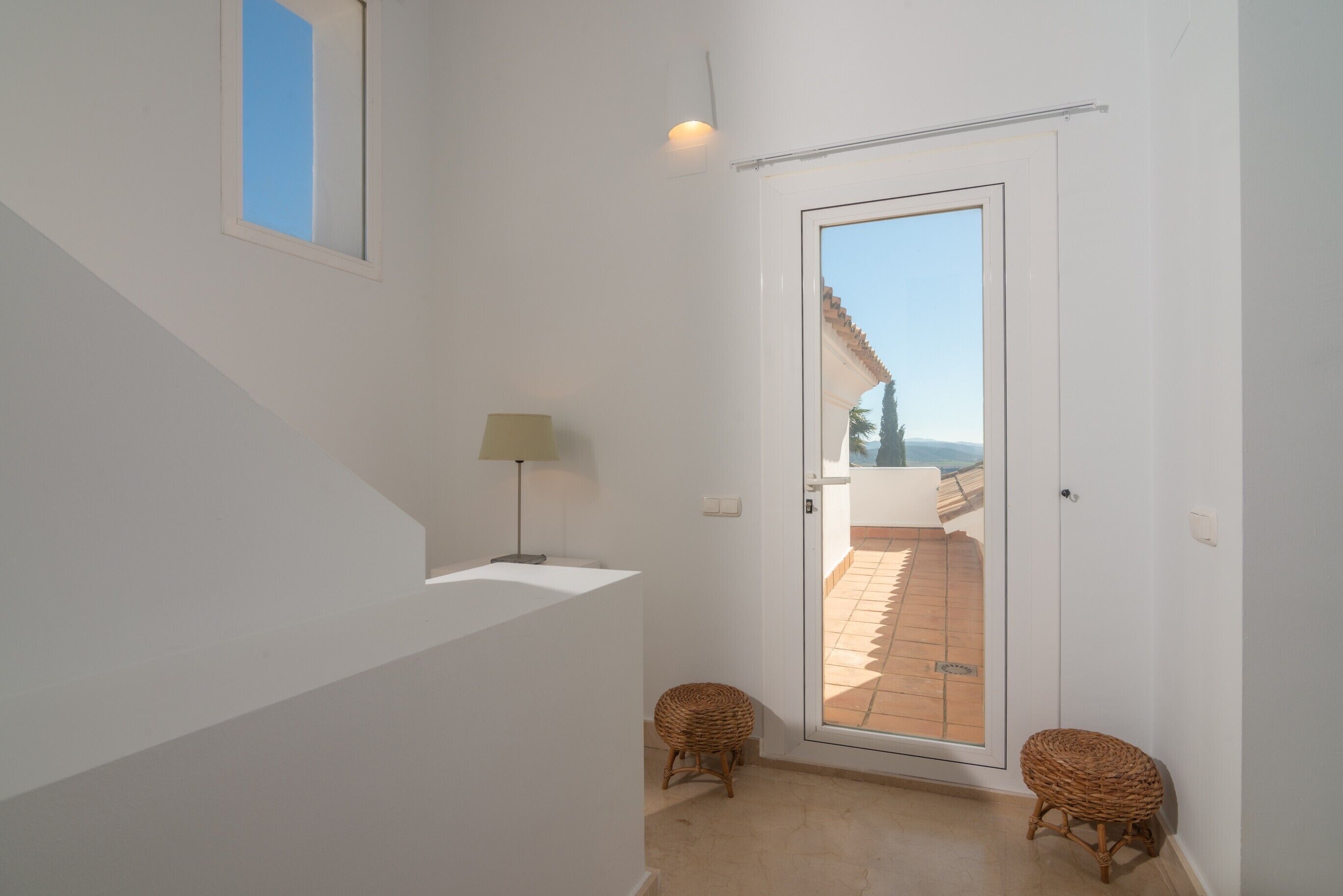 Un interior blanco con una puerta que da a un espacio exterior, una lámpara y dos taburetes de mimbre.