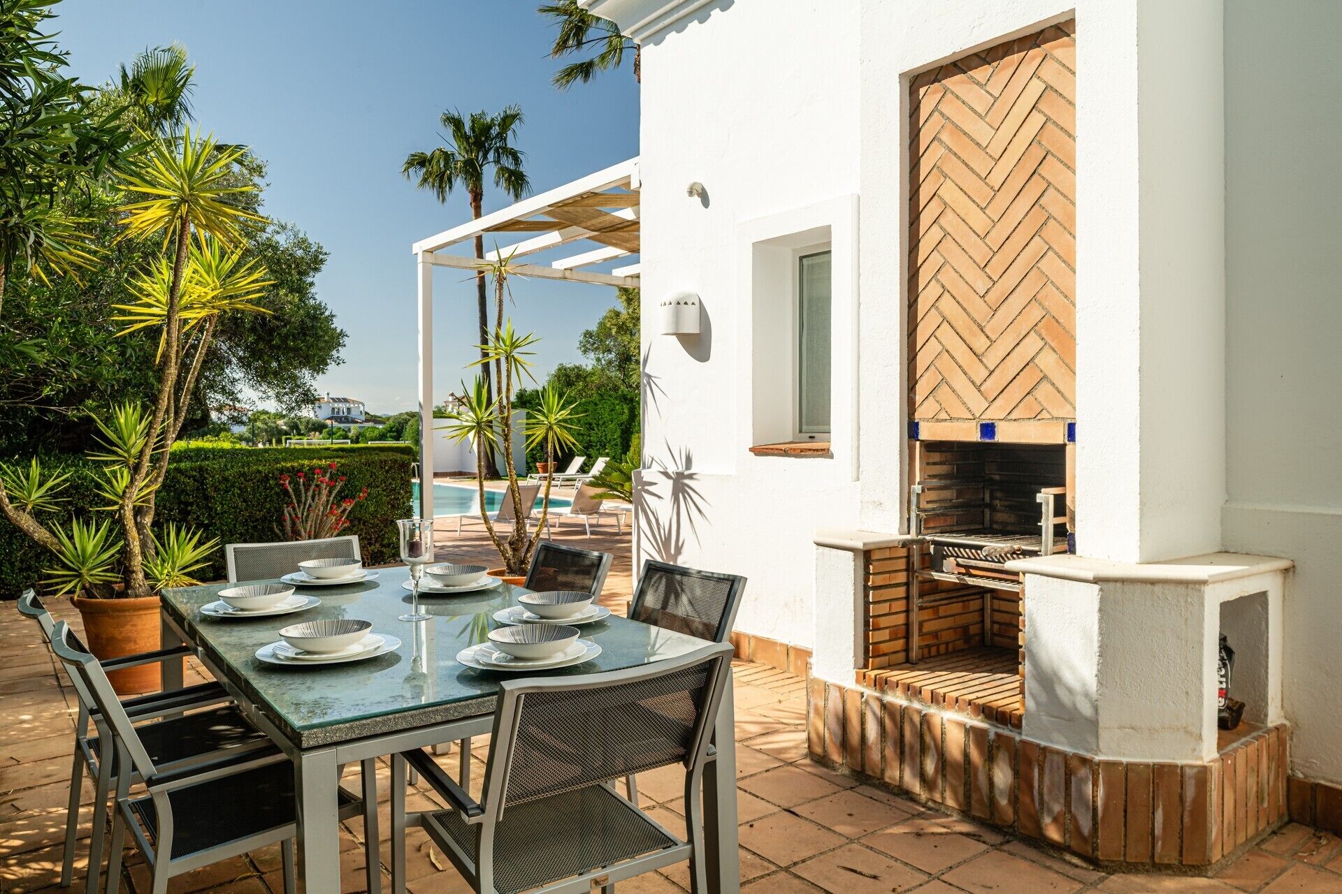 Een terras met eettafel voor vier personen, omringd door stoelen, een bakstenen barbecue en een wit gebouw met een houten deur.