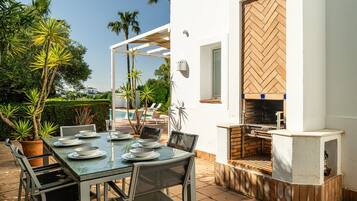 Een terras met eettafel voor vier personen, omringd door stoelen, een bakstenen barbecue en een wit gebouw met een houten deur.