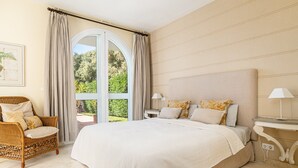 A bedroom with a large bed, a wicker chair, a nightstand with a lamp, and a view of greenery through a window. - Villa Rosa with Pool, Garden and Wi-Fi (Benalup-Casas Viejas)