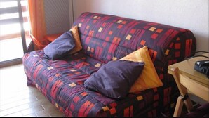A sofa with patterned upholstery and two pillows, a radiator, and a wooden chair in a room with a window. - T2 mezzanine near beach and shops (Saint-Cyprien)