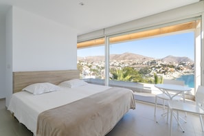 Ein Schlafzimmer mit einem großen Bett, einem kleinen Tisch und Stühlen sowie einem großen Fenster mit Blick auf eine Stadt und Berge.