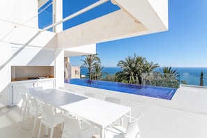 Un espace repas extérieur moderne, doté d’une piscine, de mobilier blanc et offrant une vue sur l’océan.