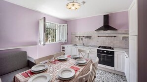 Une cuisine de taille réduite avec des meubles blancs, une cuisinière à gaz et une fenêtre agrémentée d’un rideau blanc.