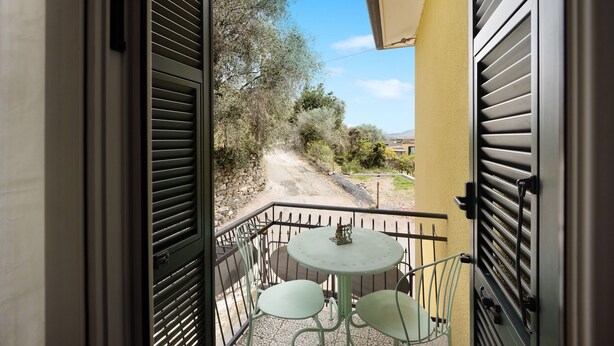 Un balcon ouvert avec une table et des chaises, donnant sur un chemin de terre et des arbres.