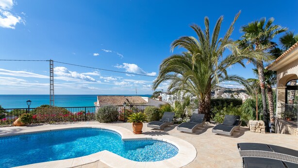 A swimming pool with lounge chairs, palm trees, and a view of the sea.