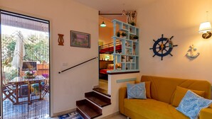 Un escalier menant à un deuxième étage, un canapé jaune orné de coussins à motifs, un gouvernail de bateau fixé au mur et une étagère avec des plantes et des objets décoratifs.